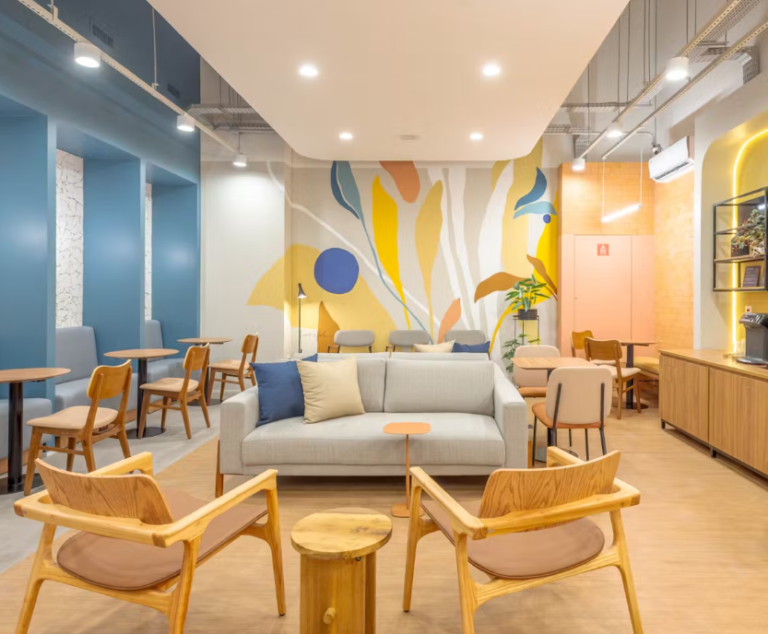 Sala iluminada com parede decorada nos tons de azul, amarelo, of white e coral, com sofás azul e cinza e cadeiras em tons de madeira clara.